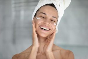 Happy excited beautiful girl enjoying facial skincare procedures, applying nourishing cream for smooth silky eye skin, massaging face with fingers, with closed eyes, smiling, laughing, Head shot